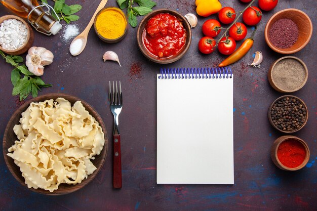 Draufsicht geschnittener roher Teig mit Tomaten und Gewürzen auf dunklem Schreibtischteig-Nudelmahlzeit-Lebensmittelgemüse