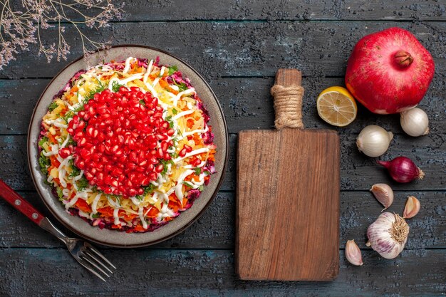 Draufsicht Granatapfel-Zitronen-Knoblauch-Schneidebrett zwischen geschälter Granatapfel-Knoblauch-Zwiebelzitrone und Teller mit Gabel auf dem Tisch