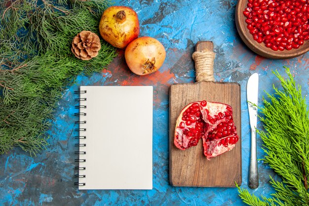 Draufsicht Granatapfelkerne in Schüssel Abendessen Messer ein geschnittener Granatapfel auf Schneidebrett ein Notebook Äste auf blauer Oberfläche