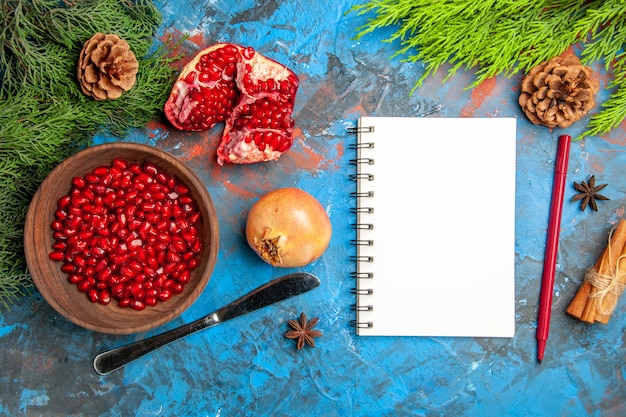 Draufsicht Granatapfelkerne in Schüssel Abendessen Messer ein geschnittener Granatapfel-Kieferzweig ein Notizbuchstift auf blauer Oberfläche