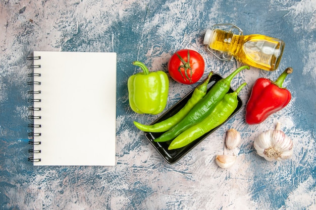 Kostenloses Foto draufsicht grüne peperoni auf schwarzem teller tomaten rote und grüne paprika knoblauch ein notizbuch auf blau-weißem hintergrund