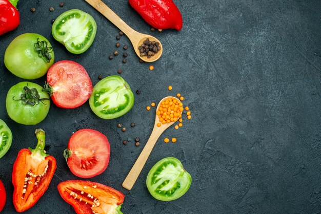 Draufsicht grüne Tomaten rote Paprika gemütliche rote Linsen und schwarzer Pfeffer in Holzlöffeln auf dunklem Tisch