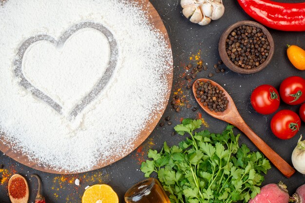 Draufsicht Herzaufdruck mit Mehl auf rundem Holzbrett schwarzer Pfeffer in kleiner Schüssel und Holzlöffel Tomaten Knoblauch Koriander auf dem Tisch