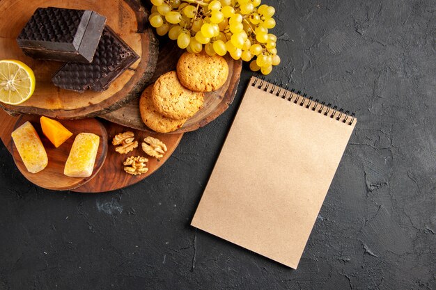 Draufsicht Holzbretter Käse geschnittene Zitronenstücke von dunklen Schokoladenkeksen Trauben Notizbuch auf dunklem Hintergrund