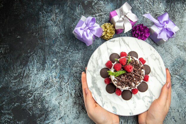 Draufsicht-Käsekuchen mit Schokolade auf ovalem Teller in den Weihnachtsgeschenken der Frauenhände auf freiem Platz der grauen Oberfläche