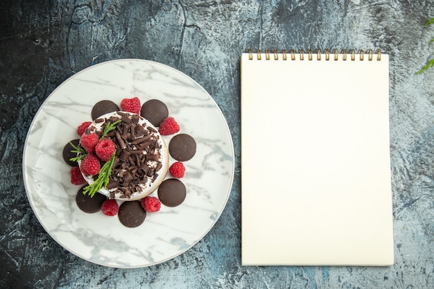 Draufsicht-Käsekuchen mit Schokolade und Himbeeren auf weißem ovalem Plattennotizblock auf grauer Oberfläche