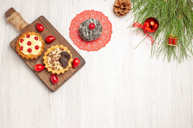 Draufsicht Kakao- und Beerentörtchen und Kornelkirschen auf dem Schneidebrettkakaokuchen und den Kiefernblättern mit Weihnachtsspielzeug auf dem weißen Holzgrund