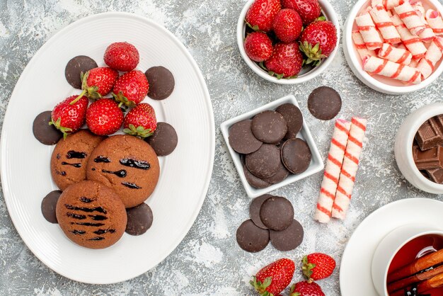 Draufsicht Kekse Erdbeeren und runde Pralinen auf den ovalen Teller Schalen mit Süßigkeiten Erdbeeren Pralinen und Zimttee auf dem grau-weißen Tisch
