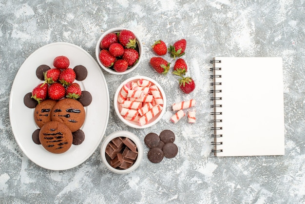 Draufsicht Kekse Erdbeeren und runde Pralinen auf den weißen ovalen Teller Schalen mit Süßigkeiten Erdbeeren Pralinen und Notizbuch auf dem grau-weißen Tisch