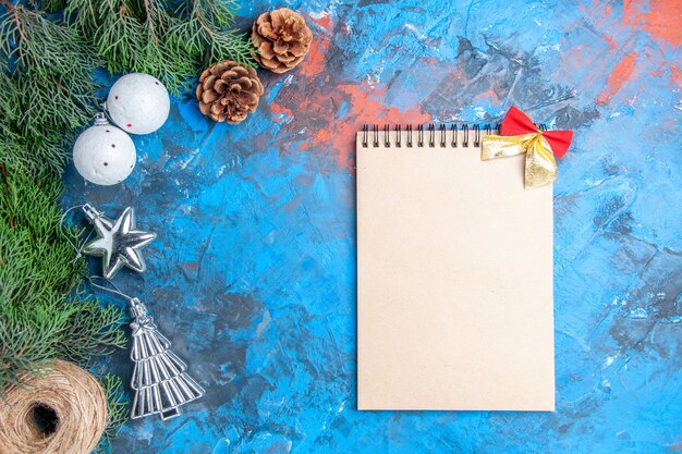 Draufsicht Kiefer Zweige Tannenzapfen Weihnachtsbaum Kugeln Stroh Faden Notizbuch mit kleiner Schleife auf blau-rotem Hintergrund