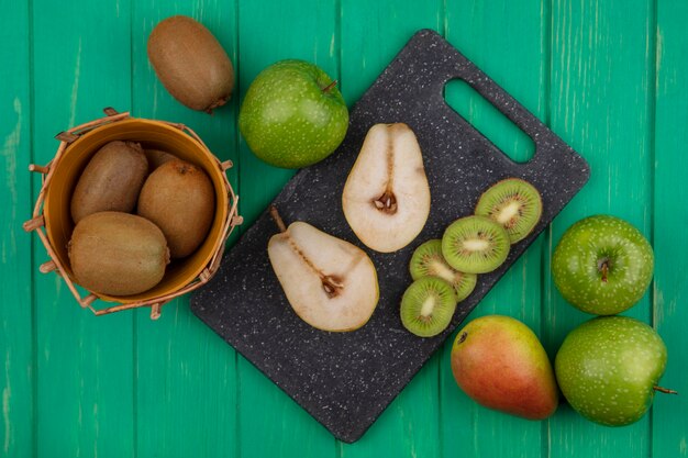 Draufsicht Kiwi in einem Korb mit grünen Äpfeln und Birnenscheiben auf einem Schneidebrett auf einem grünen Hintergrund