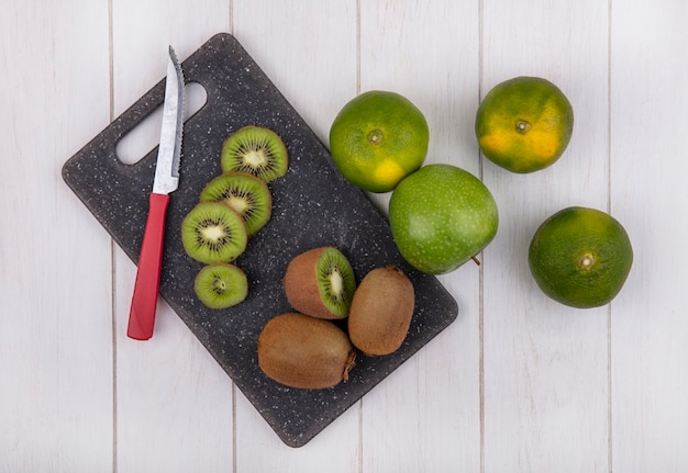 Draufsicht Kiwi mit Messer auf Schneidebrett mit Mandarinen und Apfel auf weißer Wand