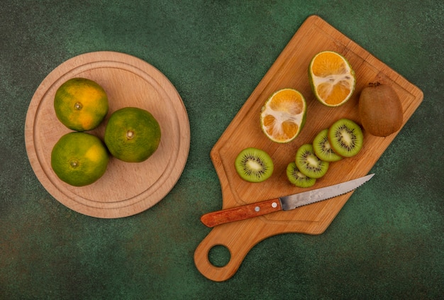 Draufsicht Kiwi-Scheiben mit einem Messer auf einem Schneidebrett mit Mandarinen auf einem Ständer an einer grünen Wand