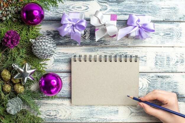 Draufsicht kleine Geschenke Weihnachtsbaum Spielzeug Tannenbaum Zweige Notizbuch Bleistift in weiblicher Hand auf Holzoberfläche