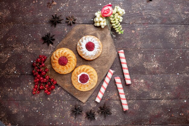 Draufsicht kleine köstliche Kuchen mit Himbeeren und frischen Preiselbeeren zusammen mit Stockbonbons auf dem braunen hölzernen Schreibtischkuchen süßer Zuckerfruchtfoto