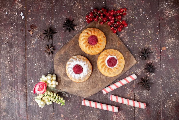 Draufsicht kleine köstliche Kuchen mit Himbeeren und frischen Preiselbeeren zusammen mit Stockbonbons auf dem braunen hölzernen Schreibtischkuchen süßes Fruchtfoto