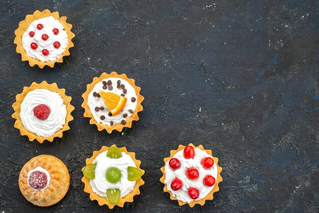 Draufsicht kleine köstliche Kuchen mit Sahne und frischen Früchten auf der dunklen Oberfläche süßer Kekskuchenzucker-Tee-Nachtisch