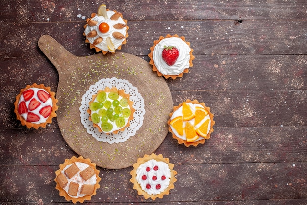 Draufsicht kleine leckere kuchen mit sahne und verschiedenen geschnittenen früchten auf dem hölzernen braunen hintergrundfruchtkuchenplätzchen süßes backfoto