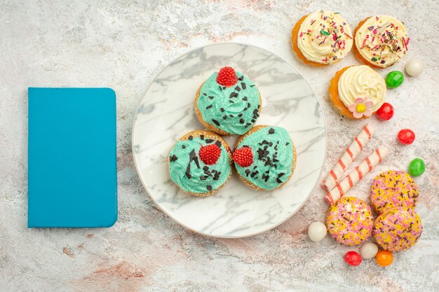 Draufsicht kleine Sahnetorten mit bunten Bonbons und Keksen auf weißer Oberfläche Leckereien Regenbogen Süßigkeiten Dessert Farbkuchen