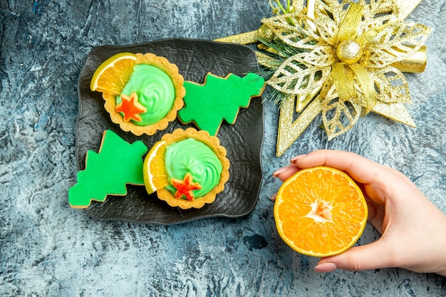 Draufsicht kleine Törtchen Weihnachtsbaumplätzchen auf schwarzem Tellerweihnachtsschmuck schneiden orange in Frauenhand auf grauem Tisch