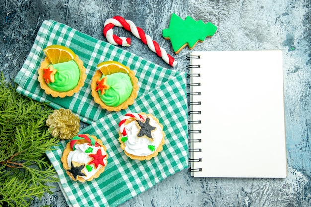 Draufsicht kleine Weihnachtstörtchen auf Tischdecke Tannenzweigen Weihnachtsbonbon-Notizblock auf grauem Tisch