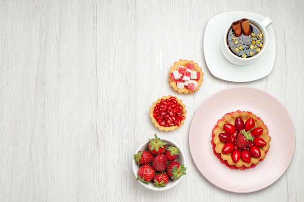 Draufsicht kleiner cremiger Kuchen mit Früchten und Tasse Tee auf weißem Schreibtisch