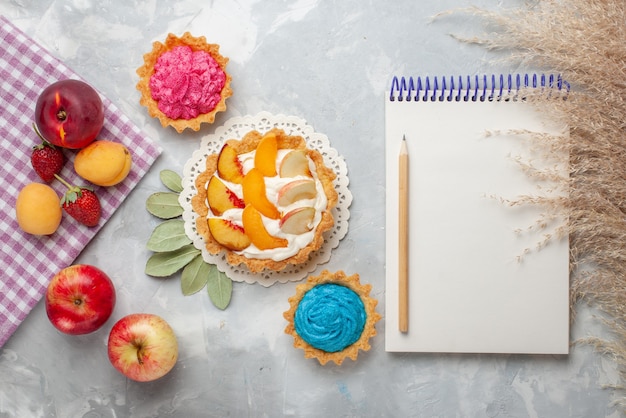 Draufsicht kleiner cremiger Kuchen mit geschnittenen Früchten und weißer Sahne zusammen mit cremigen Kuchen und Früchten auf hellweißem Schreibtischfruchtkuchenkeks süß