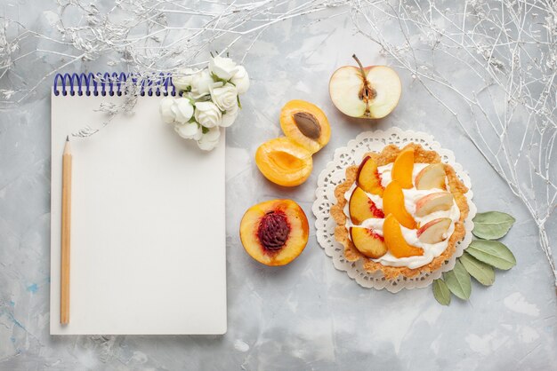 Draufsicht kleiner cremiger Kuchen mit geschnittenen Früchten und weißer Sahne zusammen mit frischem Aprikosen- und Pfirsichnotizblock auf dem weißen hellen Schreibtischfruchtkuchen-Keksplätzchen