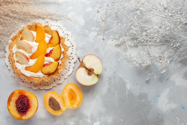 Draufsicht kleiner cremiger Kuchen mit geschnittenen Früchten und weißer Sahne zusammen mit frischen Aprikosen und Pfirsichen auf Weißlichtschreibtisch-Obstkuchen-Keksplätzchen