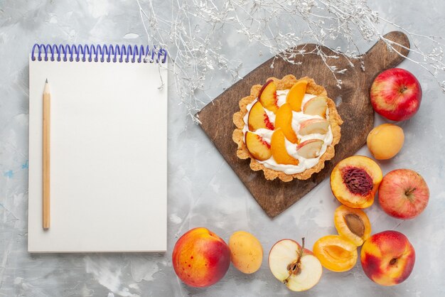 Draufsicht kleiner cremiger Kuchen mit geschnittenen Früchten und weißer Sahne zusammen mit frischen Früchten und Notizblock auf hellweißem Schreibtischfruchtkuchen-Sahneauflauf