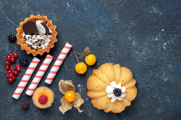 Draufsicht kleiner köstlicher Kuchen zusammen mit Keks- und rosa Stockbonbonfrüchten auf dem dunklen Schreibtischkekskuchen süße Frucht