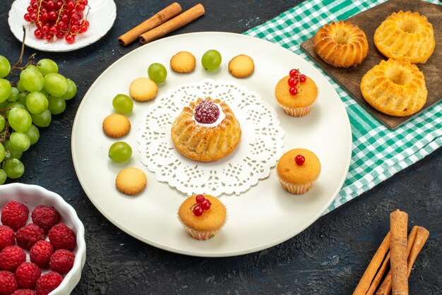 Draufsicht kleiner Kuchen mit Sahnekeksen und grünen Trauben Himbeeren und Preiselbeeren auf dem dunklen Schreibtisch süß