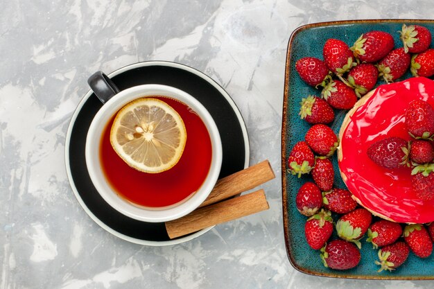 Draufsicht köstlich aussehender Kuchen kleiner Kuchen mit Tasse Tee und frischen Erdbeeren auf hellweißem Oberflächenkuchen-Keks-Kekscreme-Zuckersüß