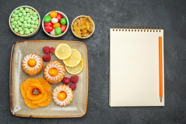 Draufsicht köstliche kleine Kuchen mit Zitronenscheiben Mandarinen und Bonbons auf dunklem Hintergrund Tee Fruchtkeks süße Kekse Pie sweet