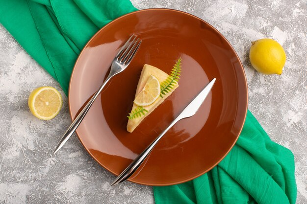 Draufsicht köstliche Kuchenscheibe mit Zitrone innerhalb brauner Platte auf dem hellen Hintergrund mit grünem Taschentuch Keksteig backen