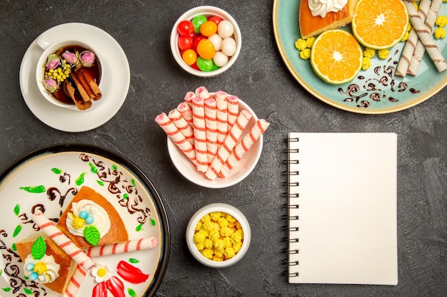 Draufsicht köstliche Tortenscheiben mit frischen Mandarinen und Tasse Tee auf dunkelgrauem Hintergrund Torte Obst Süßigkeiten Kuchen Teig Tee cake