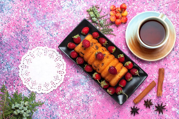 Draufsicht köstlicher gebackener Kuchen innerhalb der schwarzen Kuchenform mit frischen roten Erdbeeren und Tee auf dem rosa Schreibtisch.