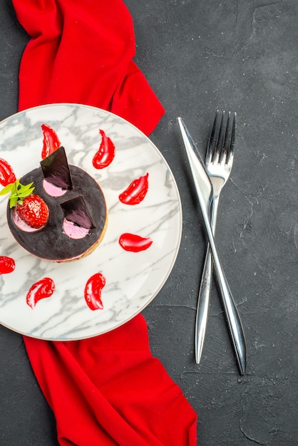 Draufsicht köstlicher Käsekuchen mit Erdbeere und Schokolade auf Teller mit rotem Schal, gekreuztem Messer und Gabel auf dunklem, isoliertem Hintergrund