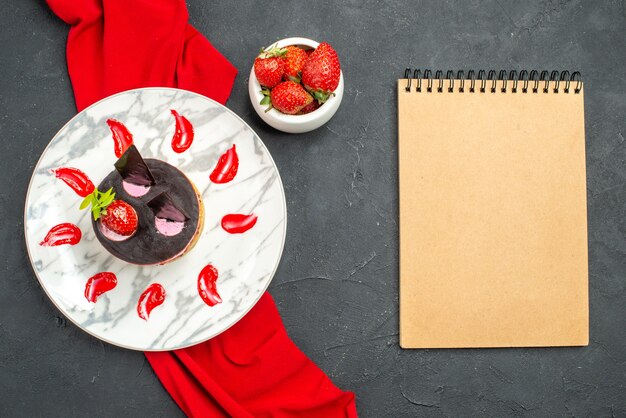 Draufsicht köstlicher Käsekuchen mit Erdbeere und Schokolade auf Teller roter Schalschale mit Erdbeeren ein Notizbuch auf dunklem, isoliertem Hintergrund