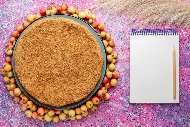 Draufsicht köstlicher runder Kuchen innerhalb Platte mit gefütterten Süßkirschen und Notizblock auf hellrosa Schreibtischkuchenkuchen-Keks süßer Backzucker