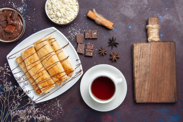 Draufsicht köstliches süßes Gebäck mit Schokolade und Tasse Tee auf dunklem Schreibtisch