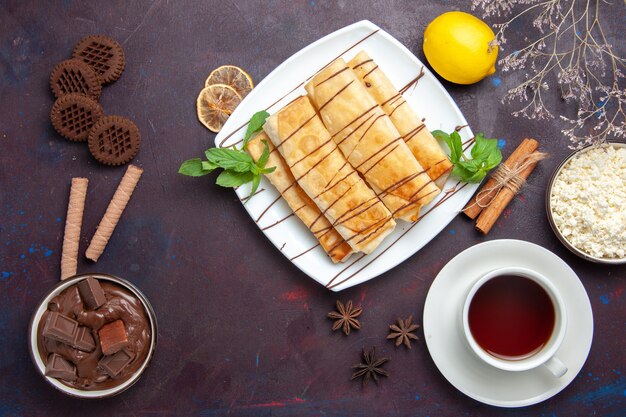 Draufsicht köstliches süßes Gebäck mit Tasse Tee und Schokolade auf dem dunklen Raum