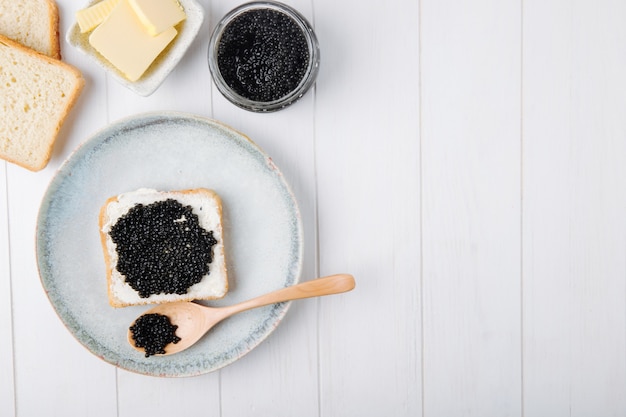 Draufsicht kopieren Raum Toast mit schwarzem Kaviar auf einem Teller mit einem Löffel und Butter mit einer Dose schwarzen Kaviars auf einem weißen Hintergrund