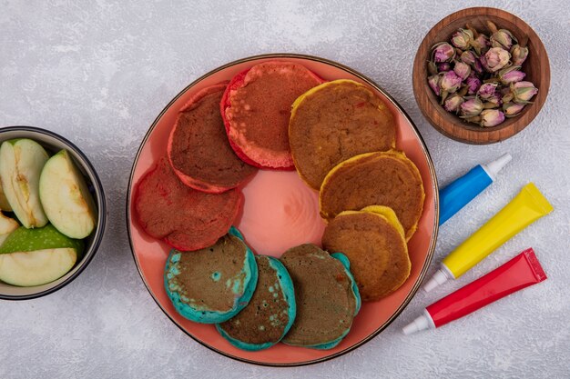 Draufsicht mehrfarbige Pfannkuchen auf einem orangefarbenen Teller mit Scheiben von grünem Apfel in einer Schüssel mit mehrfarbigen Farben in Röhren auf einem weißen Hintergrund