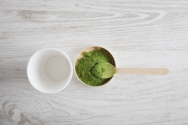 Draufsicht nehmen weißes Papierglas und Premium-Bio-Japan-Matcha-Tee auf Holztisch weg, bereit für moderne Weise Lattezubereitung. Präsentation erster Schritt. Teelöffel grünes Pulver nehmen.