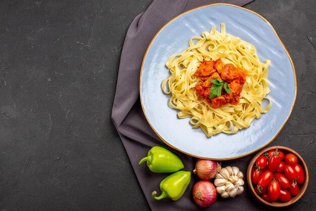 Draufsicht Nudeln auf Tischdecke Teller mit appetitlichen Nudeln neben der Schüssel Tomaten Knoblauch Zwiebel grüner Kugelpfeffer auf der lila Tischdecke auf dem Tisch