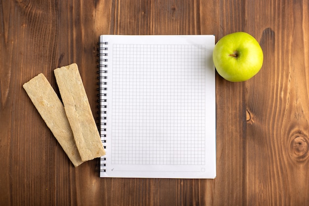 Draufsicht offenes blaues Heft mit Crackern und grünem Apfel auf dem braunen Schreibtisch