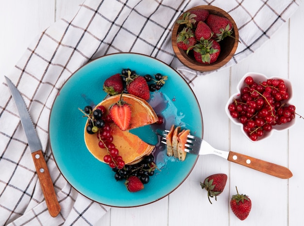 Draufsicht Pfannkuchen mit Erdbeeren der schwarzen und roten Johannisbeeren mit einer Gabel und einem Messer auf einem Teller auf einem weißen karierten Handtuch