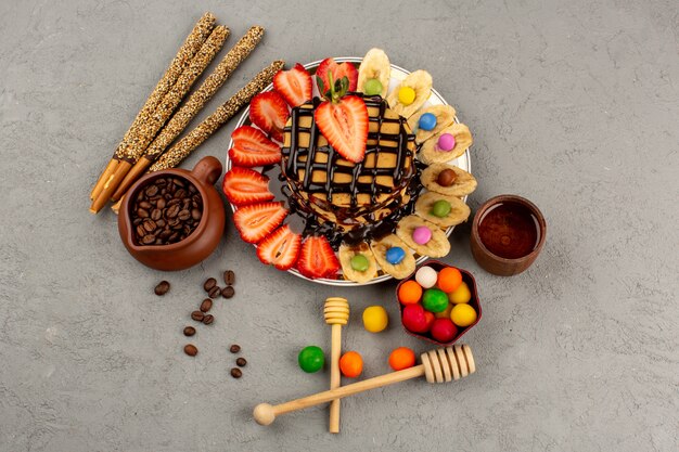 Draufsicht Pfannkuchen mit schokoladenrot geschnittenen Erdbeeren und Bananen zusammen mit Zuckerstangen und bunten Süßigkeiten auf dem grauen Boden