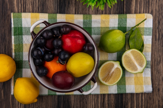 Draufsicht Pfirsich mit Orangenpflaume und Kirschpflaume in einem Topf mit Limetten und Zitronen auf einem karierten Handtuch auf einem hölzernen Hintergrund